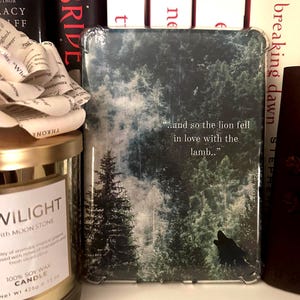 Puede incluir: Una funda protectora transparente para lector de libros electrónicos con un diseño de bosque y el texto "...and so the lion fell in love with the lamb." La funda tiene una silueta de lobo negro. Se ven libros y una vela.