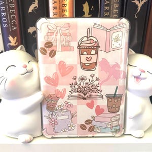 May include: A pink and white chequered phone case with illustrations of coffee cups, books, hearts, flowers, and coffee beans. The case has a clear border and is flanked by two white cat figurines.