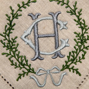 May include: A white linen napkin with a green embroidered wreath and a blue embroidered monogram "H" in the center. A blue bow is at the bottom of the wreath.