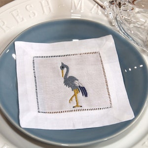 May include: White linen napkin with a blue and yellow embroidered heron design. The napkin is folded and placed on a blue plate.