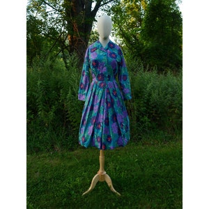 Può includere: Un abito vintage a fiori con sfondo blu e fiori viola e rossi. L'abito ha un colletto, maniche lunghe e una gonna plissettata.