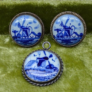 May include: A set of vintage jewelry featuring a pendant and matching earrings. Each piece has a round, blue and white ceramic design depicting a windmill scene, set in a silver-toned metal frame. The earrings are stud style.