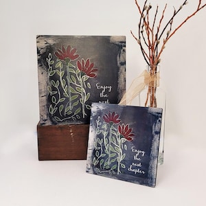 May include: Two square decorative plaques with a dark blue background and a floral design. The flowers are red with green stems and leaves. The text "Enjoy the next chapter" is written on the plaques. A vase with branches is in the background.