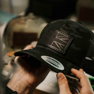 Op de afbeelding: Een zwarte en groene camouflage baseballpet met een zwart leren patch met de Union Jack-vlag. De pet heeft een zwart mesh achterpaneel en een wit label met de tekst "The Authentic Snapback".