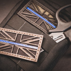 Op de afbeelding: Twee zwarte en bruine stoffen patches met een blauwe lijn en een Union Jack-ontwerp zijn bevestigd aan een zwarte tactische tas. Een paar zwarte tactische scharen zijn ook bevestigd aan de tas.