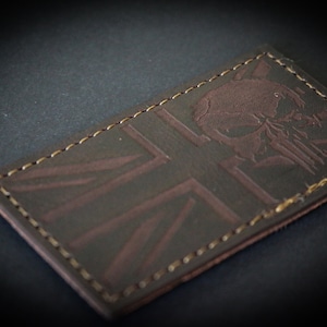 May include: A brown leather patch with a Union Jack design and a skull emblem. The patch has a stitched border with gold thread. The skull is embossed on the upper right corner. The patch is rectangular in shape.