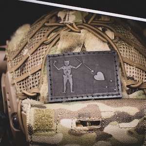 Puede incluir: Un parche negro y blanco con una calavera y huesos cruzados, un corazón y una flecha. El parche está unido a un casco de camuflaje verde y beige.