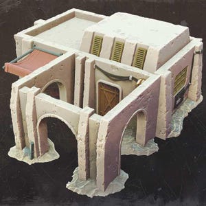 Może przedstawiać: Szczegółowy model miniaturowy budynku w stylu pustynnym z płaskim dachem, łukami i czerwonym markizą. Budynek ma beżowo-czerwonobrązową kolorystykę, z detalami architektonicznymi takimi jak okna i drzwi.