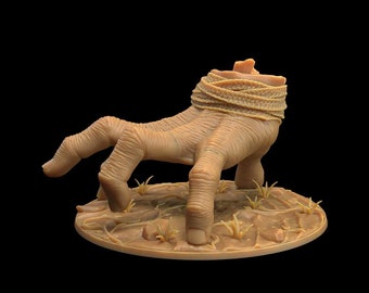 Pode incluir: Uma miniatura detalhada pintada à mão de uma mão esquelética que emerge do solo. A mão está envolta em bandagens e tem uma aparência texturizada e realista. A miniatura está colocada sobre uma base com uma superfície texturizada e pequenos elementos marrons que parecem grama.