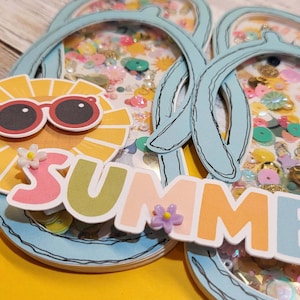 Könnte beinhalten: Eine sommerliche Karte mit einem blauen Flip-Flop-Design. Die Karte hat ein transparentes Fenster, das mit bunten Pailletten und Glitzer gefüllt ist. Das Wort "SUMMER" ist in bunten Buchstaben geschrieben, mit einer gelben Sonne, die eine Sonnenbrille trägt, oben drauf.
