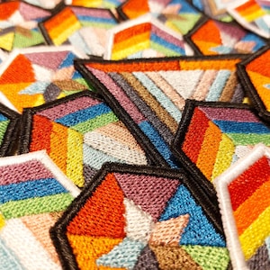 May include: A close-up of a collection of colorful embroidered patches. The patches are hexagonal in shape and feature a geometric design with a rainbow color scheme.