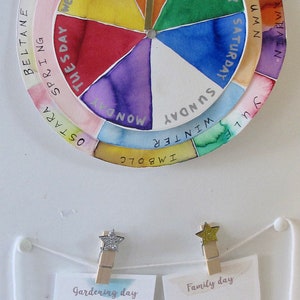 K&ouml;nnte beinhalten: Ein kreisf&ouml;rmiger Wandkalender mit einem drehbaren Zeiger, der die Wochentage in der Mitte und jahreszeitliche Feste auf dem &auml;u&szlig;eren Ring anzeigt. Darunter h&auml;ngen zwei Karten an einer Schnur mit der Aufschrift Gardening day und Family day.