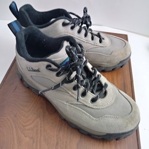 May include: A pair of gray suede and canvas hiking shoes with black laces and a black and blue trim. The shoes are on a wooden surface.