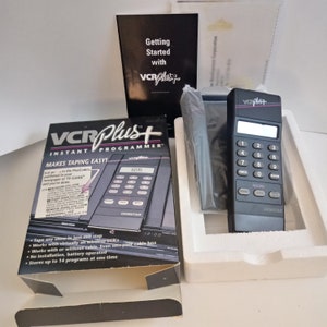 May include: A black VCR Plus Instant Programmer device with a digital display and a keypad. The device is in its original packaging, which features the product name and a description of its features. The packaging is white with black text and a black and white image of the device.
