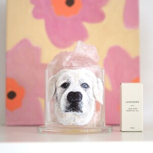 May include: A white dog's face is painted on a clear glass candle jar with a pink crystal on top. The jar is sitting on a white shelf next to a small bottle labelled "Lavender 100% Pure Essential Oil".