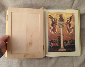 May include: An open book with a wooden cover and a page showing a religious painting of three figures in a golden frame. The figures are standing on a column with a golden background.