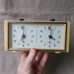 May include: A yellow plastic dual-face analogue timer with black hands and red second hands. The timer has a white face with black numbers and the word "Jantar" printed on the top. The timer is made in the USSR.