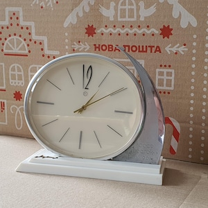 Puede incluir: Un reloj de escritorio vintage plateado y blanco con una esfera redonda y agujas negras. El reloj tiene un diseño de media luna en el lateral y el texto "CCCP BOCTOK 12 IV 1961" en la base.