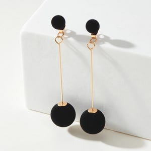May include: A pair of drop earrings featuring a small black sphere at the top and a larger black sphere at the bottom, connected by a thin gold-toned metal bar. The design includes a gold-toned jump ring connecting the top sphere to the vertical bar.