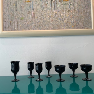 May include: Seven black glass wine glasses on a teal surface. The glasses are all different shapes and sizes.