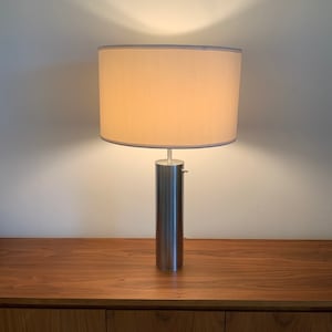 May include: A silver table lamp with a white drum shade. The lamp is on a wooden surface.
