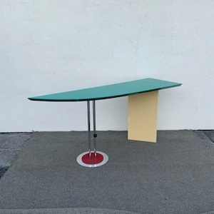 May include: A green glass-top desk with a curved edge and a yellow and white base. The desk has a chrome and red metal base with a round foot.