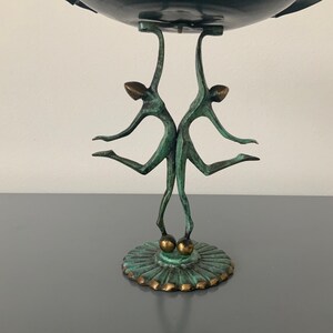 May include: A green bronze sculpture of two figures holding up a bowl. The figures are standing on a round base with a decorative design.