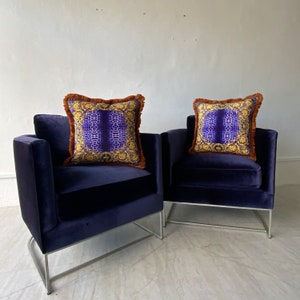 May include: Two navy blue velvet armchairs with silver metal legs. Each chair has a decorative square pillow with a purple and gold pattern and a brown fringe. The chairs are placed against a white wall.