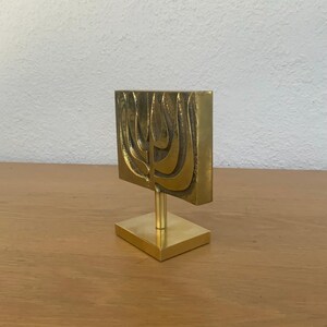 May include: A gold-toned metal sculpture of a menorah on a square base. The menorah is stylized and abstract, with a textured surface.