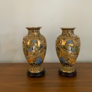 May include: A pair of gold and black ceramic vases with intricate hand-painted designs featuring figures in traditional Japanese clothing. The vases have a round body and a flared lip.