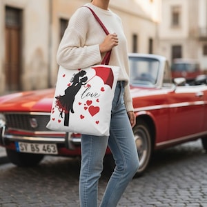 以下が含まれることがあります： 赤い持ち手が付いた白いトートバッグ。赤い傘の下にいるカップルの黒いシルエット、"love"の文字と赤いハートが描かれています。白いセーターとブルージーンズを着た人がバッグを持っています。