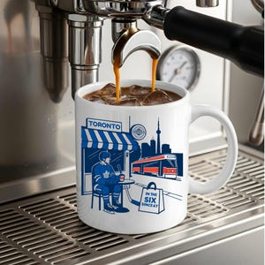 May include: White ceramic mug with a Toronto-themed graphic. The design features a hockey player, a streetcar, and the CN Tower in blue and red. Text on the mug reads "TORONTO" and "IN THE SIX SINCE 67". Coffee is being poured into the mug.