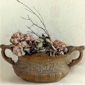 Könnte beinhalten: Eine antike, bronzefarbene Zierurne mit zwei Griffen, gefüllt mit getrockneten Hortensien und Zweigen. Die Urne hat ein geprägtes Blumendesign vor einem neutralen Hintergrund.
