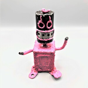 May include: A pink robot sculpture with a black head and a silver face. The robot has two arms and three legs. The robot is made of metal and has a textured surface.