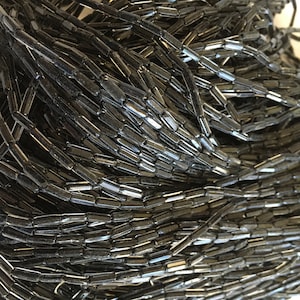 Op de afbeelding: Een stapel zwarte glazen kralen, elke kraal is een lange, dunne cilinder.