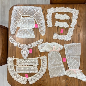 May include: An assortment of antique white lace garments, including a collar, sleeves, and a bodice. The delicate lace pieces are arranged on a wooden table, showcasing intricate floral patterns and designs. The collection appears to be vintage, with a focus on delicate craftsmanship.