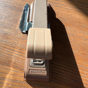 May include: A tan and brown Bostitch stapler. The stapler has a silver metal handle and a tan plastic base. The word "BOSTITCH" is printed on the front of the stapler. The stapler is sitting on a wooden surface.