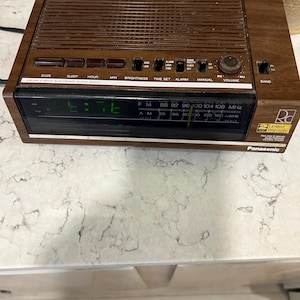 May include: A vintage Panasonic FM-AM electronic clock radio with a dark wood grain finish. The digital display shows the time. Features include snooze, sleep, and alarm functions. The radio has a volume dial and a band selector.
