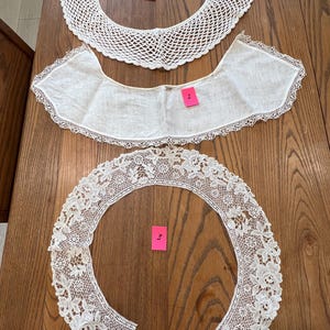 May include: Three antique white lace collars displayed on a wooden table. The collars feature different lace patterns, including a crocheted net design, a delicate scalloped edge, and floral lace detailing. Each collar is labeled with a pink tag.