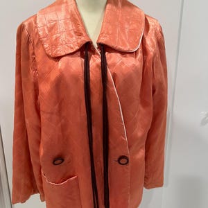 May include: A peach-colored quilted jacket featuring a wide collar, long sleeves, and two dark brown buttons on the front. The garment includes two front pockets and two long, thin dark brown fabric ties hanging from the neckline area.