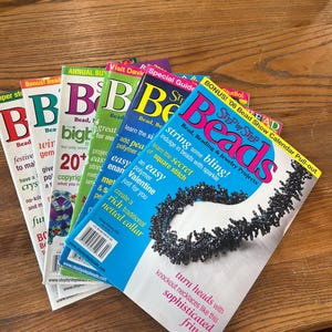 Könnte beinhalten: Ein Stapel farbenfroher "Beads"-Magazine mit verschiedenen Titeln, darunter "Bonus! Bead" und "Step by Step Beads". Die Zeitschriften zeigen Bilder von Perlenschmuck und Texte wie "string some bling!" und "knockout necklaces".