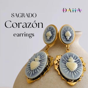 May include: Gold-toned earrings with a light blue oval pendant featuring a cream-colored heart design. The earrings have a decorative gold filigree frame and are labeled "SAGRADO Corazón earrings". The earrings are displayed against a neutral background.