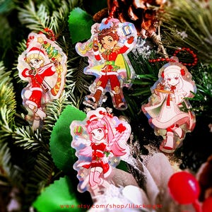 May include: Four Christmas-themed acrylic charms with cartoon characters in festive outfits. The charms are red, white, and gold, with a holographic effect. They are hanging on a background of pine needles and greenery. The text "www.etsy.com/shop/lilacknight" is visible.