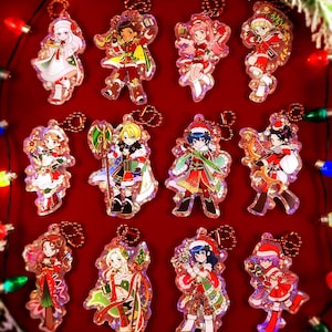 May include: A collection of Christmas-themed acrylic keychains featuring anime-style characters in festive outfits. Each keychain has a unique design with vibrant colors, attached to a metal ring. The background is a deep red with Christmas lights.