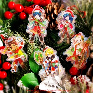 May include: A collection of five Christmas-themed acrylic charms featuring anime-style characters in festive outfits. The charms are clear with colorful designs, set against a backdrop of Christmas greenery and red berries. Each charm has a red string for hanging.