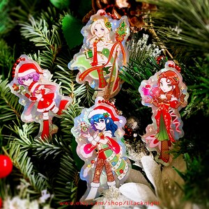 May include: Four Christmas-themed anime character acrylic charms with red, green, and white outfits and Santa hats. The charms are hanging on a Christmas tree with green needles and red berries.