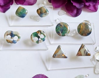 May include: A collection of six pairs of earrings in various shapes: rounded, skull-shaped, and triangular. The earrings feature a mix of colors including green, blue, gold, and white. They are displayed on clear acrylic stands, with purple flowers in the background.