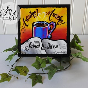 May include: A colorful painting of a coffee mug with steam rising from it, a heart shape in the steam, and the words "Awake!" on either side of the mug. The mug is sitting on top of an open book with the words "Hebrews" and "With Jesus & Java" on the pages. The painting is framed in black.
