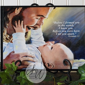 May include: A watercolor painting of Jesus holding a baby. The painting is framed in a black frame and has the text "Before I formed you in the womb, I knew you. Before you were born I set you apart." Jeremiah 1:5
