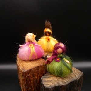 Könnte beinhalten: Drei handgefertigte Filzpuppen mit Blumenakzenten. Eine Puppe ist rosa, eine gelb und eine grün. Jede Puppe hat eine einzigartige Frisur und ist auf einem rustikalen Holzsockel vor schwarzem Hintergrund platziert.